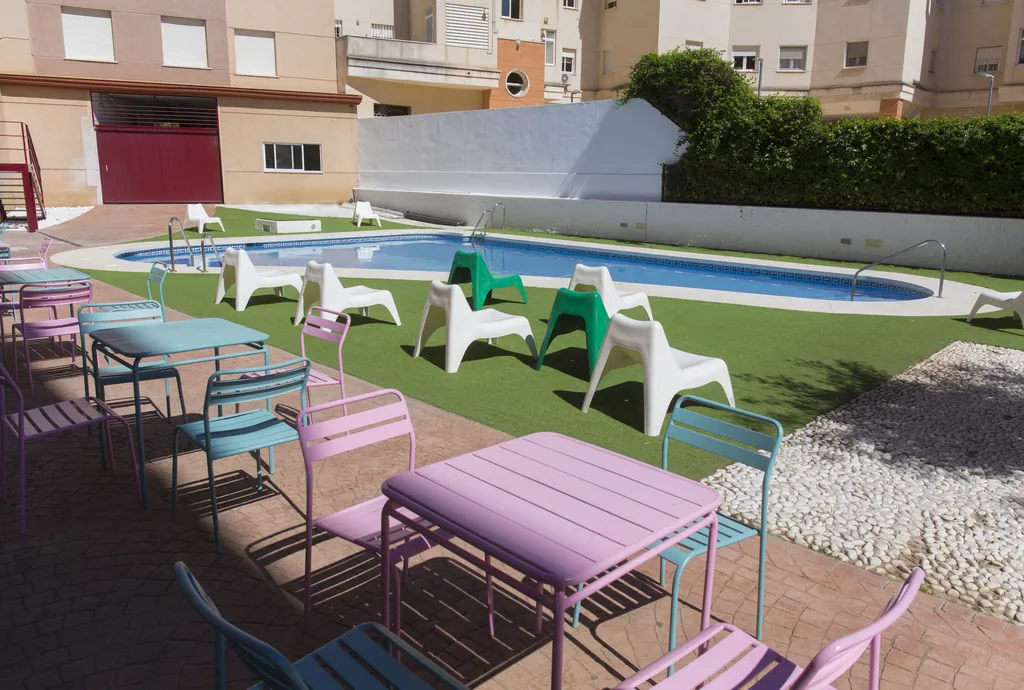 Patio de la residencia con piscina, césped y mesas de colores bajo el sol de Málaga
